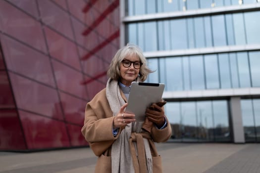 A senior woman stands outside a contemporary glass building, actively engaging with her digital tablet while dressed in a stylish brown coat and scarf on a cloudy day.