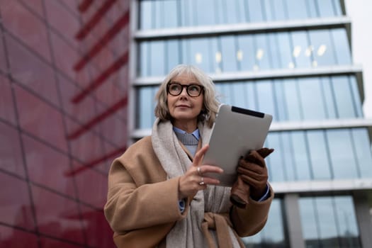 A professional woman stands outside a contemporary building, holding a tablet and dressed in a fashionable coat. She appears focused and engaged in her work.