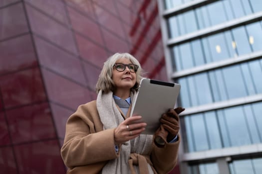 A woman with glasses and gray hair stands outside, holding a tablet. She wears a beige coat and gray scarf, focused on her device amid the striking architecture.