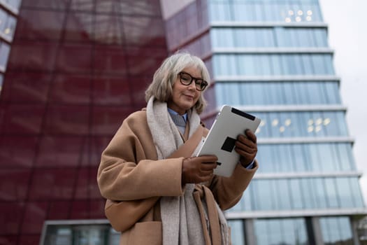 An elderly woman stands outside, dressed in a warm brown coat and scarf, comfortably using a tablet. She is surrounded by modern architectural structures on an overcast day.