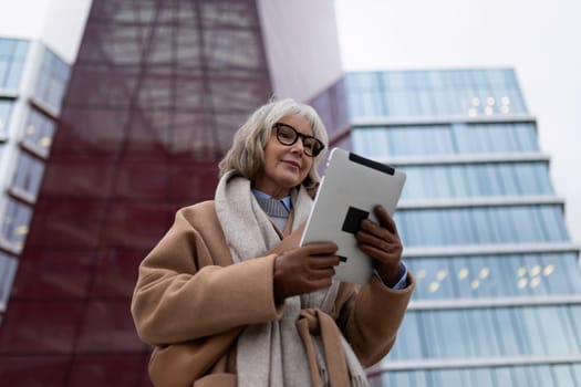 A senior woman is engaged with a tablet, standing outside contemporary buildings. The reflective glass and bold structure create a striking backdrop as twilight descends.