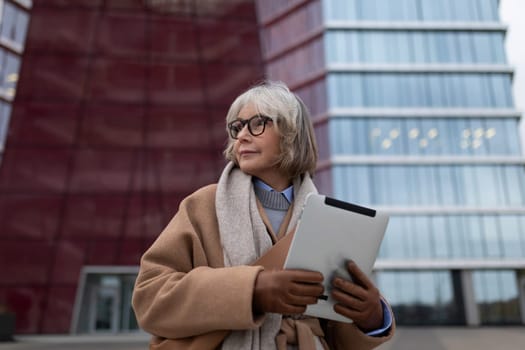 A woman with short silver hair wearing glasses stands outside in a stylish coat, holding a tablet. She looks contemplative in front of a large, contemporary building.