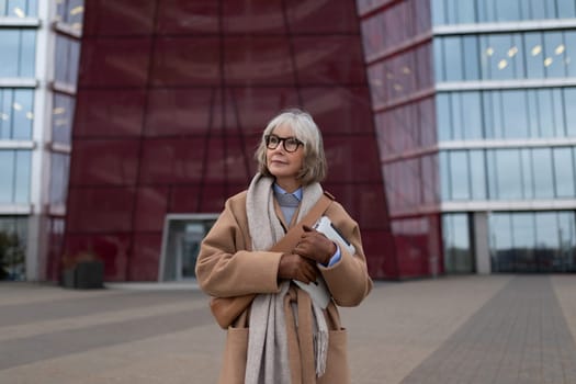 An elegant older woman holds a folder while dressed in a stylish coat and scarf, standing in front of an impressive modern building with expansive glass windows, enjoying a clear day.