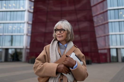 A mature woman stands confidently outside a contemporary building, holding a laptop while wearing a stylish coat and scarf. She appears thoughtful and engaged in her surroundings.