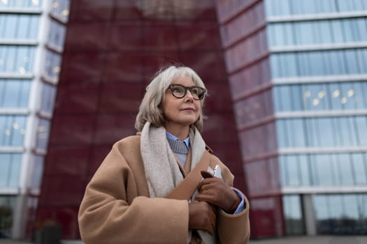A senior woman with short gray hair and glasses stands outside a striking modern structure. She wears a cozy coat and holds a bag, looking thoughtful and assured amidst the urban setting.