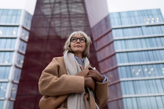 The senior woman stands gracefully with her arms crossed, dressed in a warm coat and scarf, showcasing her elegant style against a contemporary architecture backdrop during dusk.