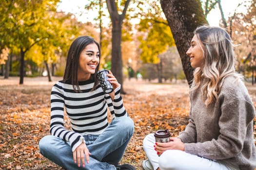 Two women are seated on the ground, engaged in conversation while holding coffee cups. They appear relaxed and comfortable in each others company.