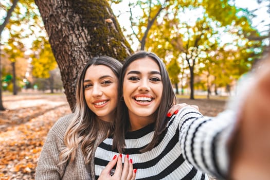 Two women, one brunette and one redhead, are smiling and posing while taking a selfie with a phone in a park. They are standing together in front of green trees under a blue sky.