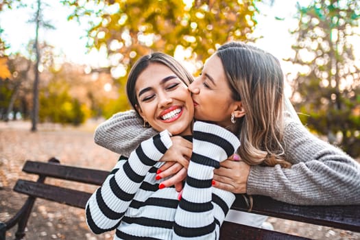 Two women of different ethnicities are kissing each other on a park bench in broad daylight. They are sitting close, leaning in towards each other, sharing an intimate moment, unaware of their surroundings.
