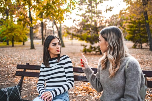 Two women are sitting on a bench in the park, angrily pointing fingers at each other and the atmosphere is heated.