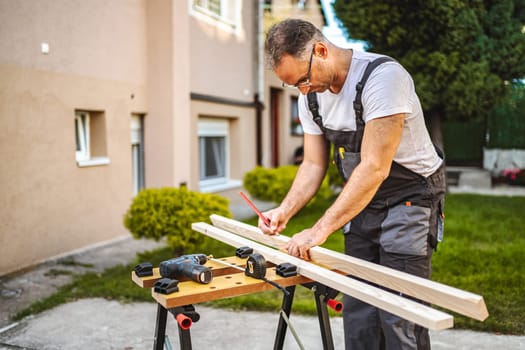 Mature male carpenter measuring wooden plank outdoors.