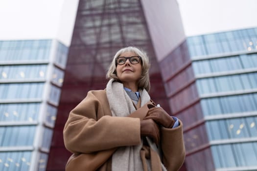 A stylish woman with gray hair and glasses poses gracefully in front of contemporary buildings, dressed warmly in a tan coat and scarf while showcasing urban elegance.
