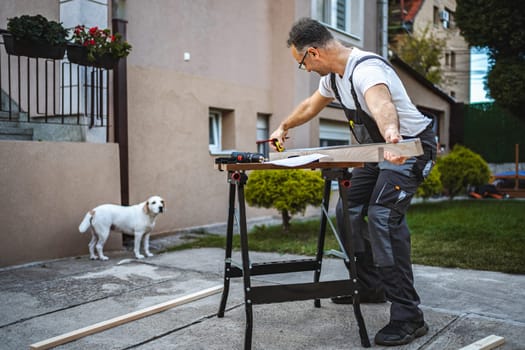 Mature male carpenter measuring wooden plank outdoors.