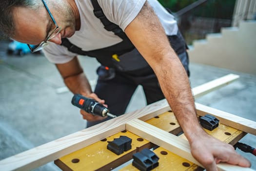 Mature male carpenter using electric screwdriver on a wooden plank outdoors.
