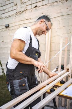 Mature male carpenter measuring wooden plank outdoors.
