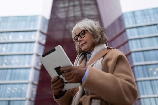 A mature woman with gray hair and glasses is focused on her tablet while standing in front of a contemporary glass structure. The scene captures an afternoon vibe.