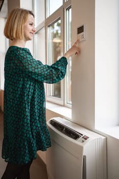 A woman wearing a green dress is standing next to a window in a room. She appears to be looking outside, with light coming in through the window. The room is simple, with minimal decor.