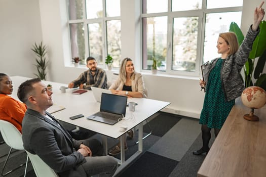 A confident woman is standing at the head of a table, presenting to her attentive colleagues in a well-lit, modern office space. Her arm is raised, likely making an important point, while her team looks on, listening and seated around the table which is equipped with a laptop, documents, and a globe.