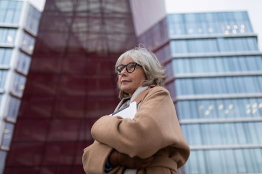 An older woman with stylish glasses and gray hair stands with her arms crossed outside a contemporary building. The urban environment showcases modern architecture and gives a lively feel.