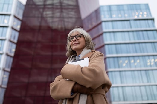 A woman with gray hair and glasses stands confidently with her arms crossed, dressed in a tan coat. Behind her is contemporary architecture reflecting the evening light.