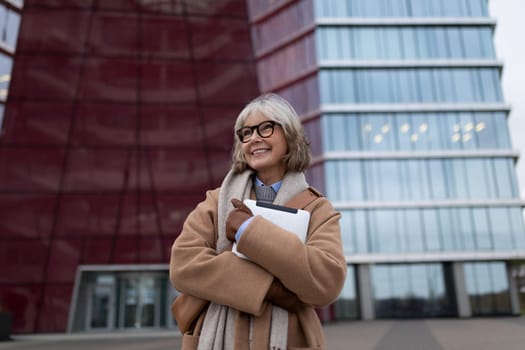 A cheerful older woman dressed in a stylish coat poses outside a sleek glass building. She holds a notebook and exudes confidence in a vibrant city environment.
