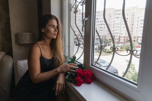 A woman in a black dress is sitting in front of a window with a bouquet of red roses