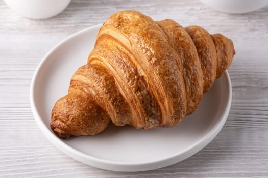 A golden, flaky croissant rests on a clean white plate, showcasing its buttery layers.