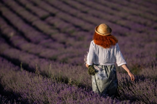Here's a caption under 150 characters describing the image: Redhead woman in straw hat walks through rows of purple lavender fields. Peaceful, scenic view.