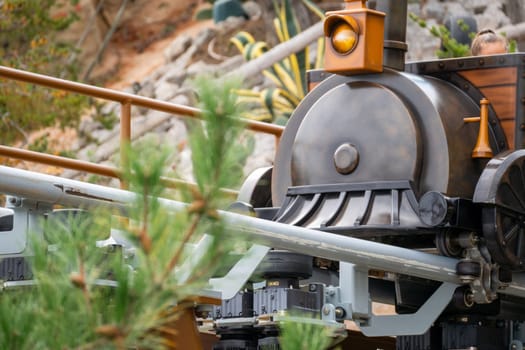 A close-up view of a black, vintage-style train engine, partially obscured by pine tree branches. The train is on a metal track.