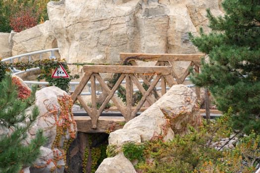 A wooden bridge spans a gap between two large, rocky outcroppings. Green foliage surrounds the bridge and rocks.
