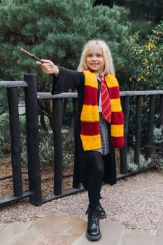 A young girl dressed as a Hogwarts student holds a wand and smiles, standing by a black fence. She wears a red and gold scarf.