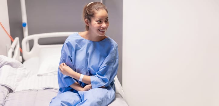 young beautiful woman in private hospital room, dressed in blue robe, sits on bed and rejoices at quick correction of cosmetic defect, waits for doctor's examination before plastic surgery,