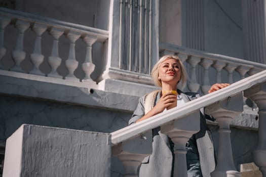 A woman is standing on a white railing with a cup of coffee in her hand. She is wearing a gray jacket and blue jeans. Concept of relaxation and leisure