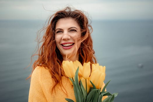 Portrait of a happy woman with hair flying in the wind against the backdrop of mountains and sea. Holding a bouquet of yellow tulips in her hands, wearing a yellow sweater.