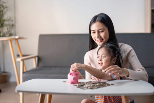 Asian mother guides her daughter in saving practice with a pink piggy bank, highlighting nurturing and learning in a home setting.