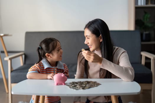 Asian mother and daughter share a sweet moment saving coins together with a piggy bank, fostering financial education in a cozy home environment.