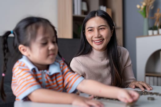 Asian mother smiles proudly watching her daughter sort coins, encouraging financial education and bonding at home with a pink piggy bank