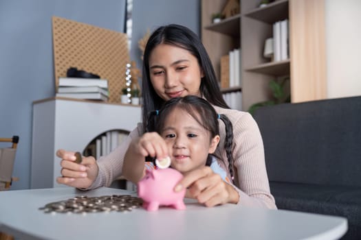 Asian mother and daughter engage in a money-saving activity using a pink piggy bank at their home, promoting learning and bonding.