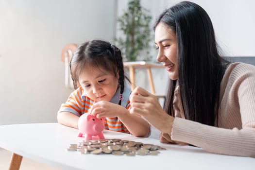 Asian mother supports her daughter in saving practice using a pink piggy bank, fostering a nurturing educational experience at home.