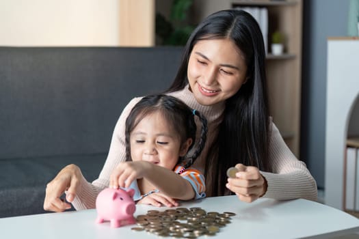 Asian mother and daughter happily save coins in a pink piggy bank at home, experiencing a joyful educational moment together.