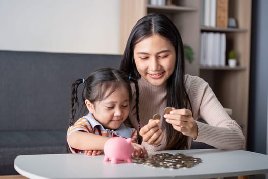 Asian mother and daughter share a bonding moment counting coins and saving them in a pink piggy bank, enhancing learning at home.
