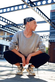A man is squatting outside in a casual outfit. He wears a grey sweatshirt, black pants, a black bandana, and white sneakers.