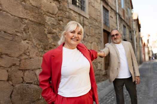 A cheerful couple walks hand in hand along a picturesque cobblestone street. The woman, dressed in red, beams with joy as they explore the historic architecture around them.