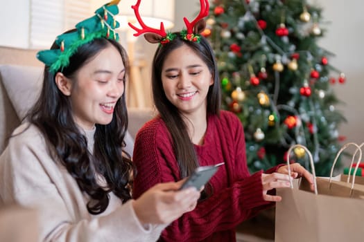 Lesbian Asian couple joyfully engaging in online shopping next to a Christmas tree, sharing smiles and holiday happiness