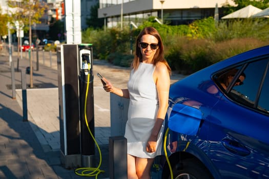 Woman in sunglasses and a fashionable white dress is using her smartphone while charging an electric car in the street.