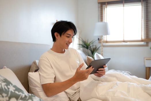 Happy adult asian man surfing internet on digital tablet in bedroom.
