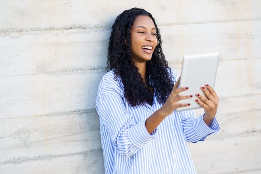 A happy woman smiles brightly while using her tablet against a modern wall, embodying the joy of technology and communication in enhancing her contemporary interactions and connectivity
