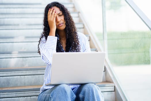 A young woman sits on stairs, overwhelmed and stressed, fixated on her laptop screen. Her expressions reveal frustration and anxiety, highlighting the pressures of modern life and studying