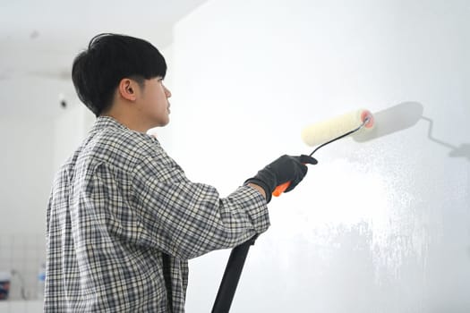Side view of a man in plaid shirt painting a wall with roller brush. Home improvement concept.