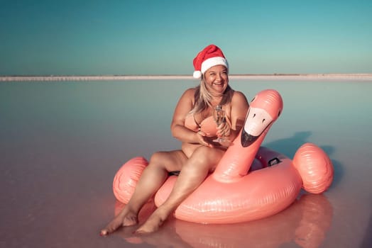 A woman wearing a Santa hat sits on a pink flamingo float in a body of water. He is smiling. There are white posts in the background.